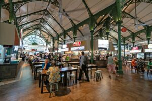 A bustling food market with ornate green iron beams. People sit at wooden tables and order from brightly lit stalls, creating a lively, social atmosphere.