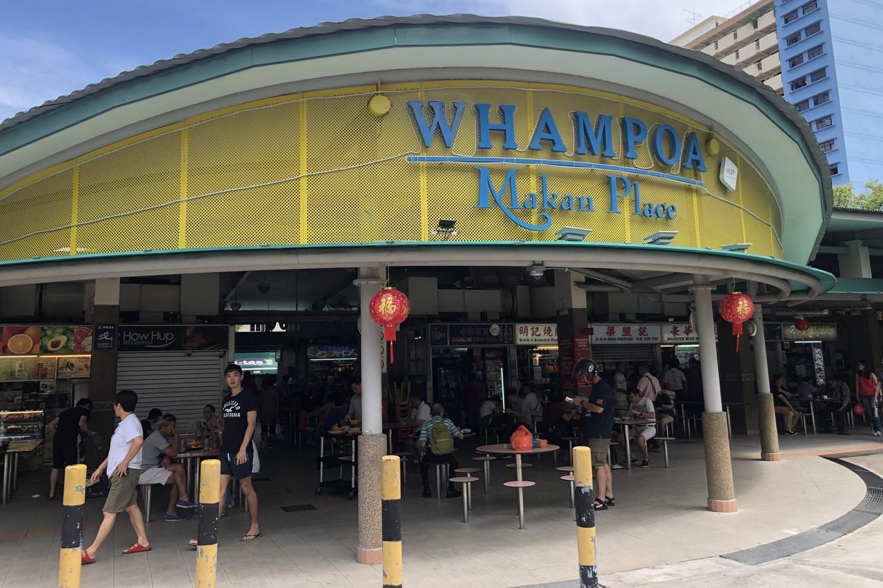  This image features the entrance to Whampoa Makan Place, marked by a curved, yellow mesh facade and blue lettering. Below the signage, people are seen walking and dining at round tables under a sheltered area decorated with red Chinese lanterns.