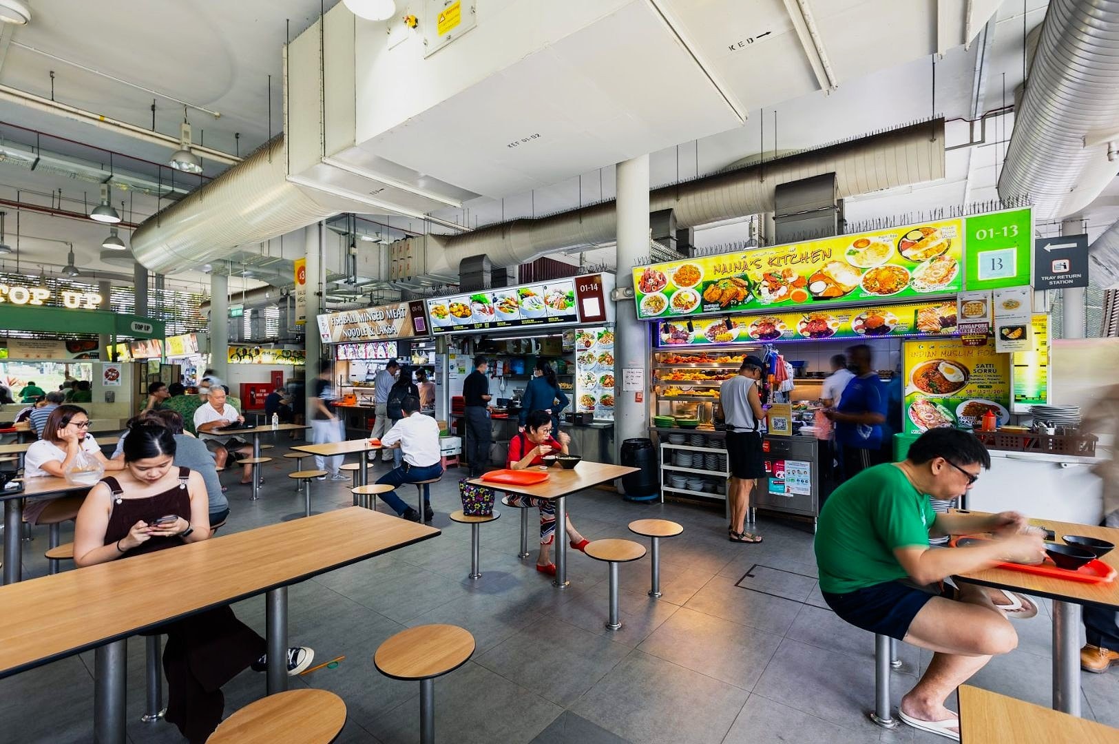 A bustling food court with colorful signage and food stalls. People sit eating or using their phones, creating a lively and casual atmosphere.