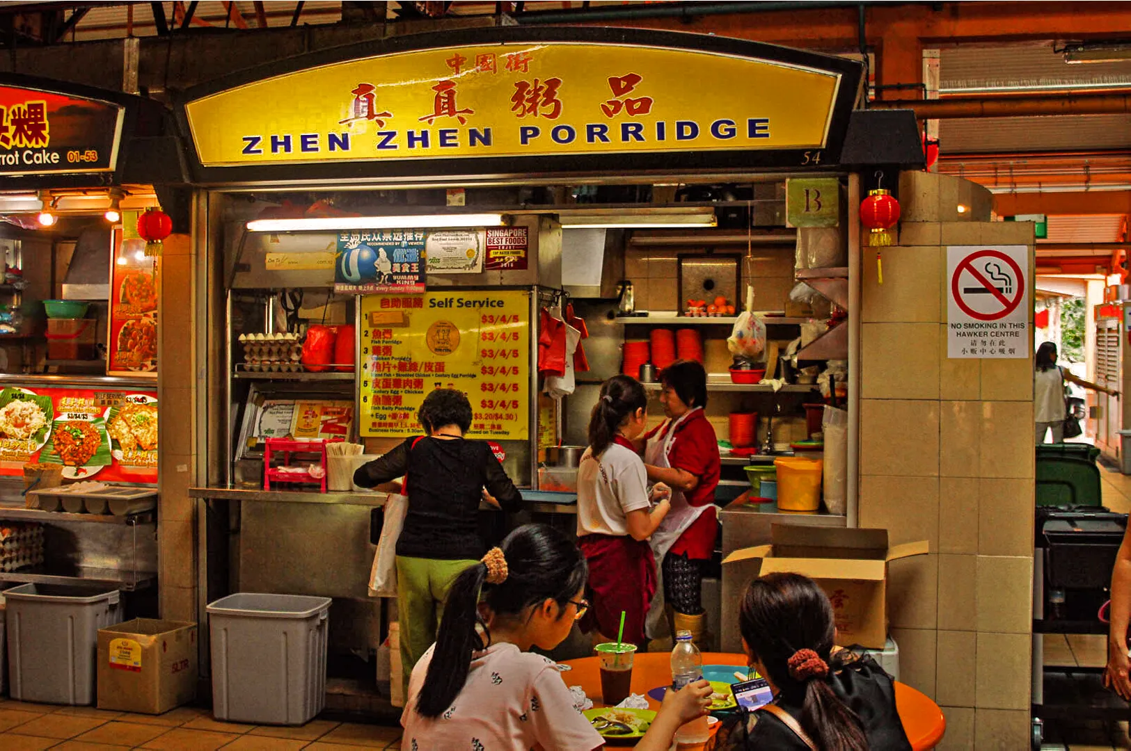 Outdoor food stall, "Zhen Zhen Porridge," in Singapore. Patrons order porridge, interacting with stall staff. Busy, lively atmosphere with a no-smoking sign.