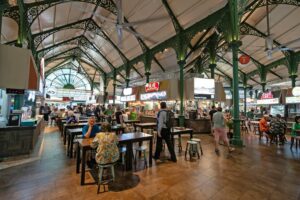 This image captures the bustling interior of Lau Pa Sat, a historic food center in Singapore, featuring its iconic green Victorian-style cast-iron structure and vaulted ceilings. People are seen dining at long wooden tables and strolling past various food stalls that offer a wide array of local cuisines.