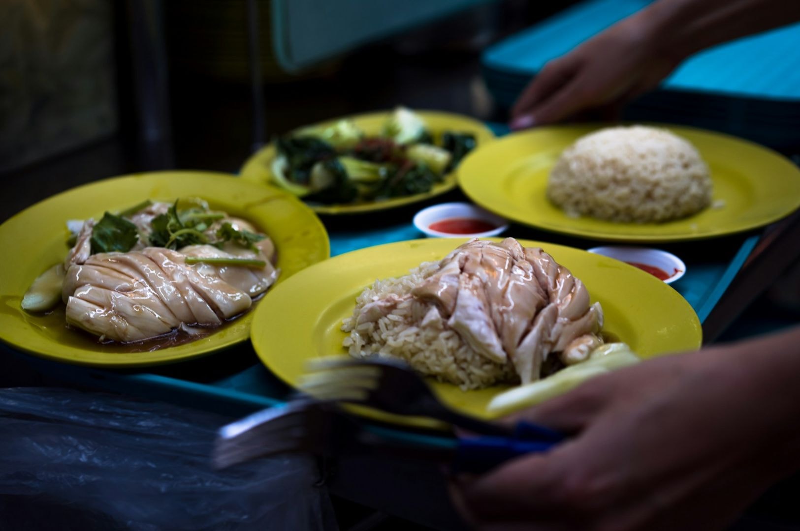 Yellow plates with chicken rice, greens, and chili sauce on a table. A person holds a plate, evoking a street food atmosphere.