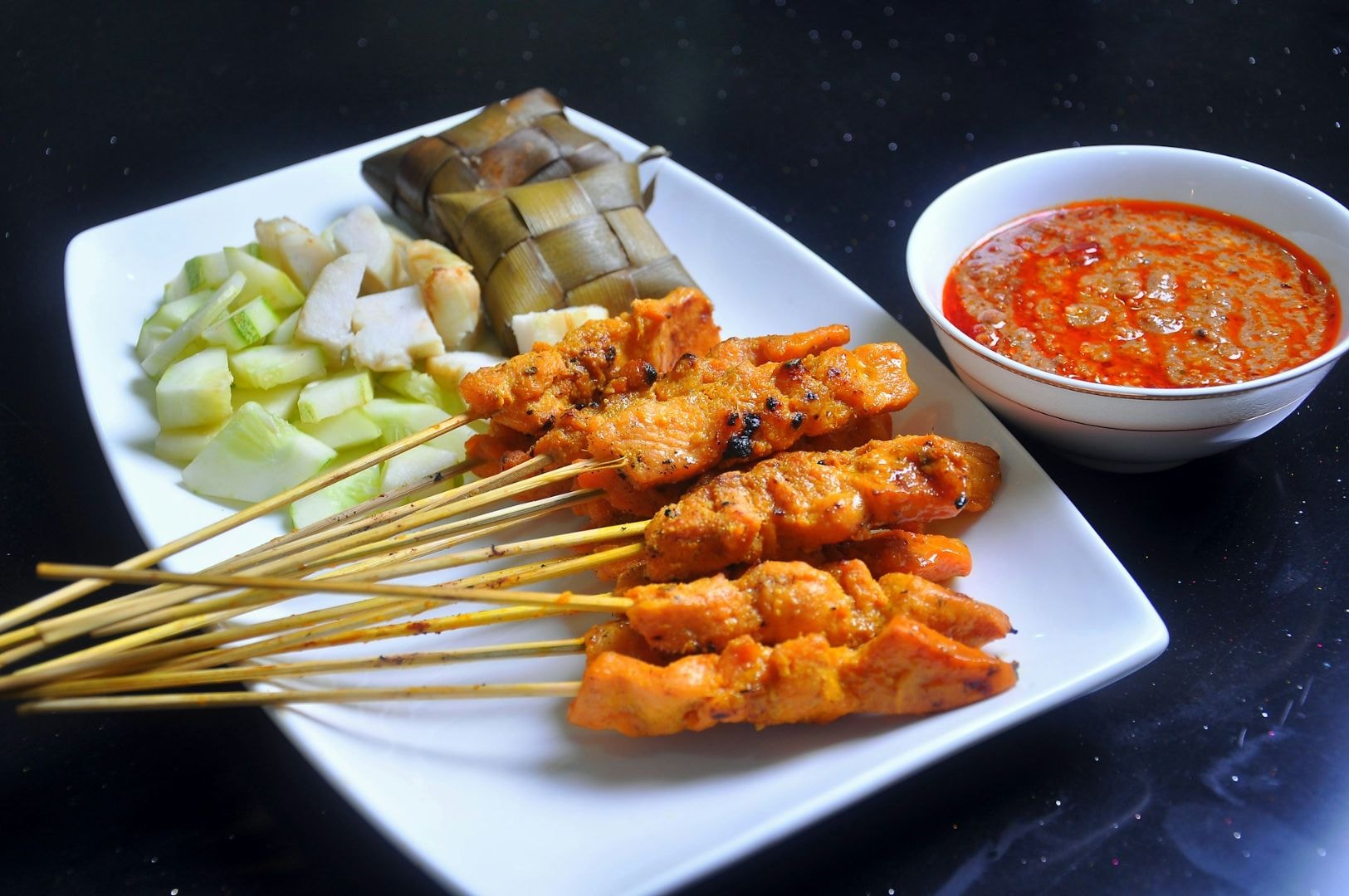 A plate of chicken satay skewers with cucumber and rice cakes, served alongside a bowl of spicy peanut sauce. The dish looks flavorful and appetizing.