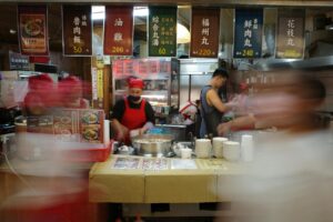 This long-exposure photograph captures the bustling interior of a Taiwanese food stall, where staff members in red aprons appear as motion-blurred figures against a backdrop of steaming pots and stacked bowls. Brightly colored hanging banners display the menu in Chinese characters, featuring traditional items like braised pork rice and meatball soup with their corresponding prices.