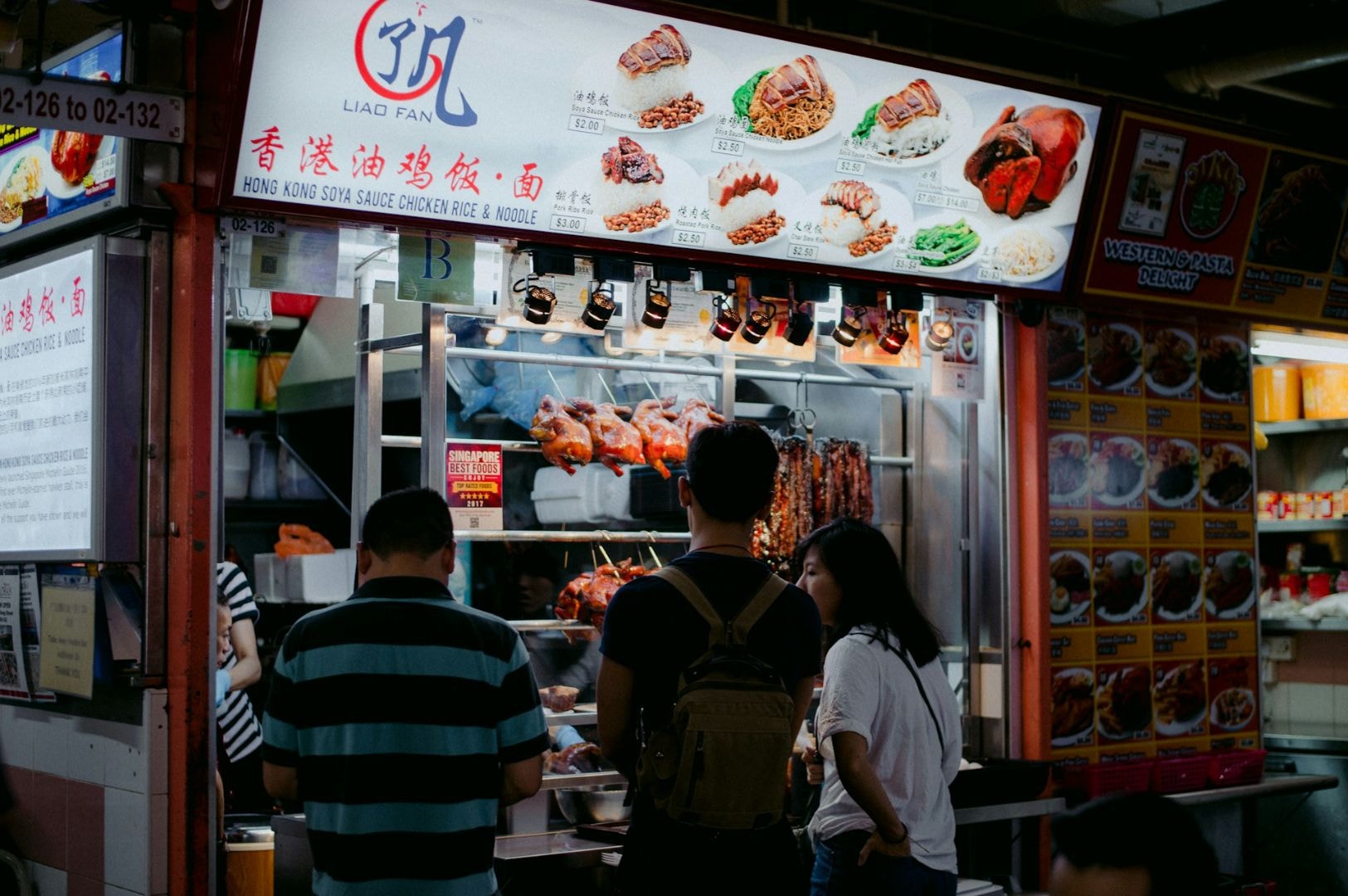 People stand at a bustling food stall featuring hanging roasted meats. A lit menu above displays Asian dishes, creating a vibrant, aromatic scene.