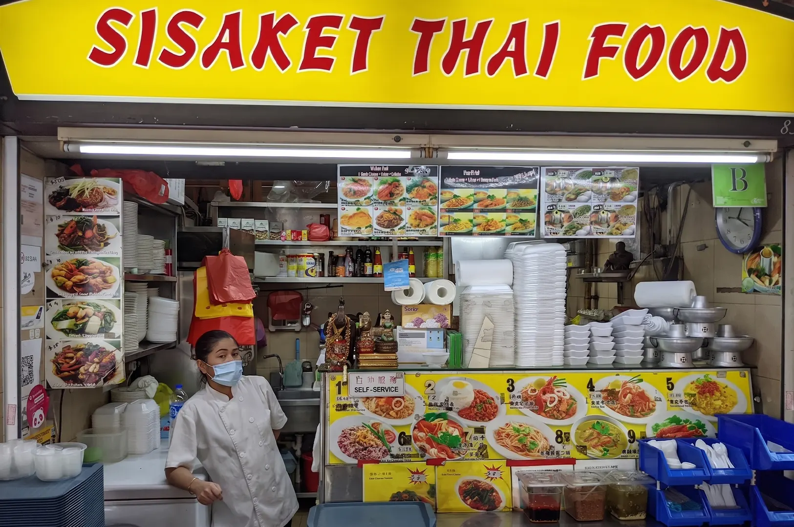 Sisaket Thai Food hawker stall storefront featuring a bright yellow sign, illustrated menu panels, and a staff member wearing a face mask.