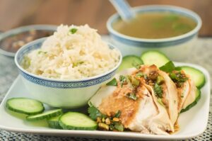 Plate of Hainanese chicken drizzled with peanut sauce, accompanied by fluffy white rice and garnished with herbs.