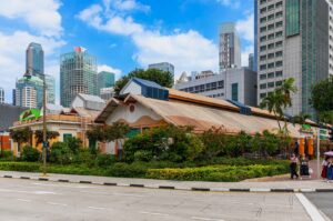 A historic market building with a wooden roof and lush greenery stands amidst modern skyscrapers. People walk on the sidewalk, conveying urban life.