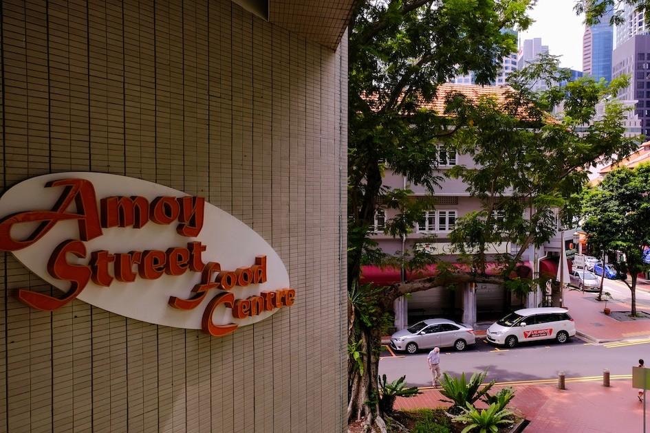  The image shows the Amoy Street Food Centre signage mounted on a textured tan wall in the foreground. Looking past the building, the scene reveals a street with parked cars, lush green trees, and traditional shophouses set against a backdrop of modern skyscrapers.