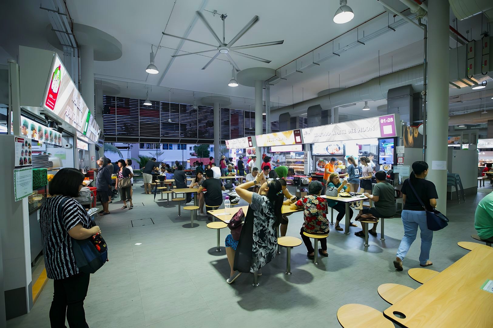 A bustling food court with people seated at tables enjoying meals, surrounded by brightly lit food stalls. The atmosphere is lively and social.