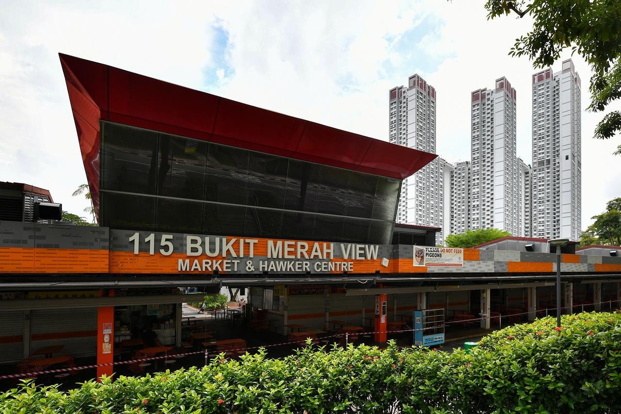  The image shows the exterior of the 115 Bukit Merah View Market & Hawker Centre in Singapore, featuring a modern glass facade with a striking red angular roofline. In the background, three tall white residential apartment blocks rise into a cloudy sky, while green hedges and red caution tape are visible in the foreground.