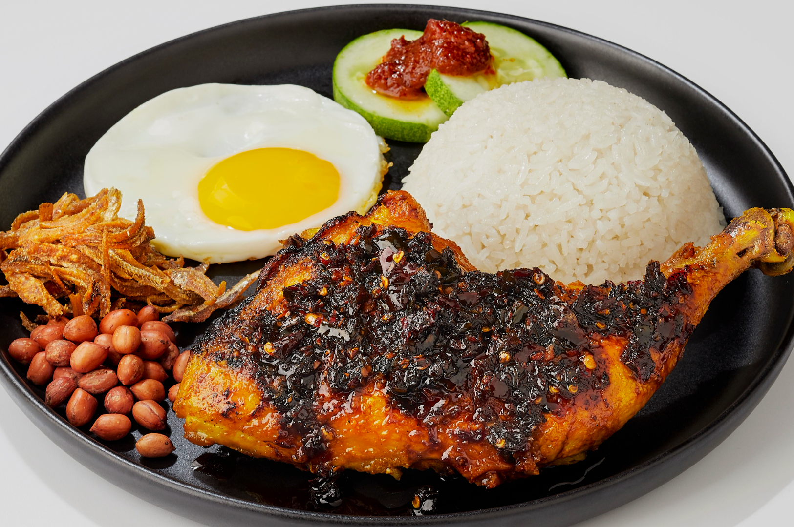 A vibrant plate of nasi lemak: sambal-coated chicken, white rice, sunny-side egg, cucumber slices with sambal, crispy anchovies, and peanuts.