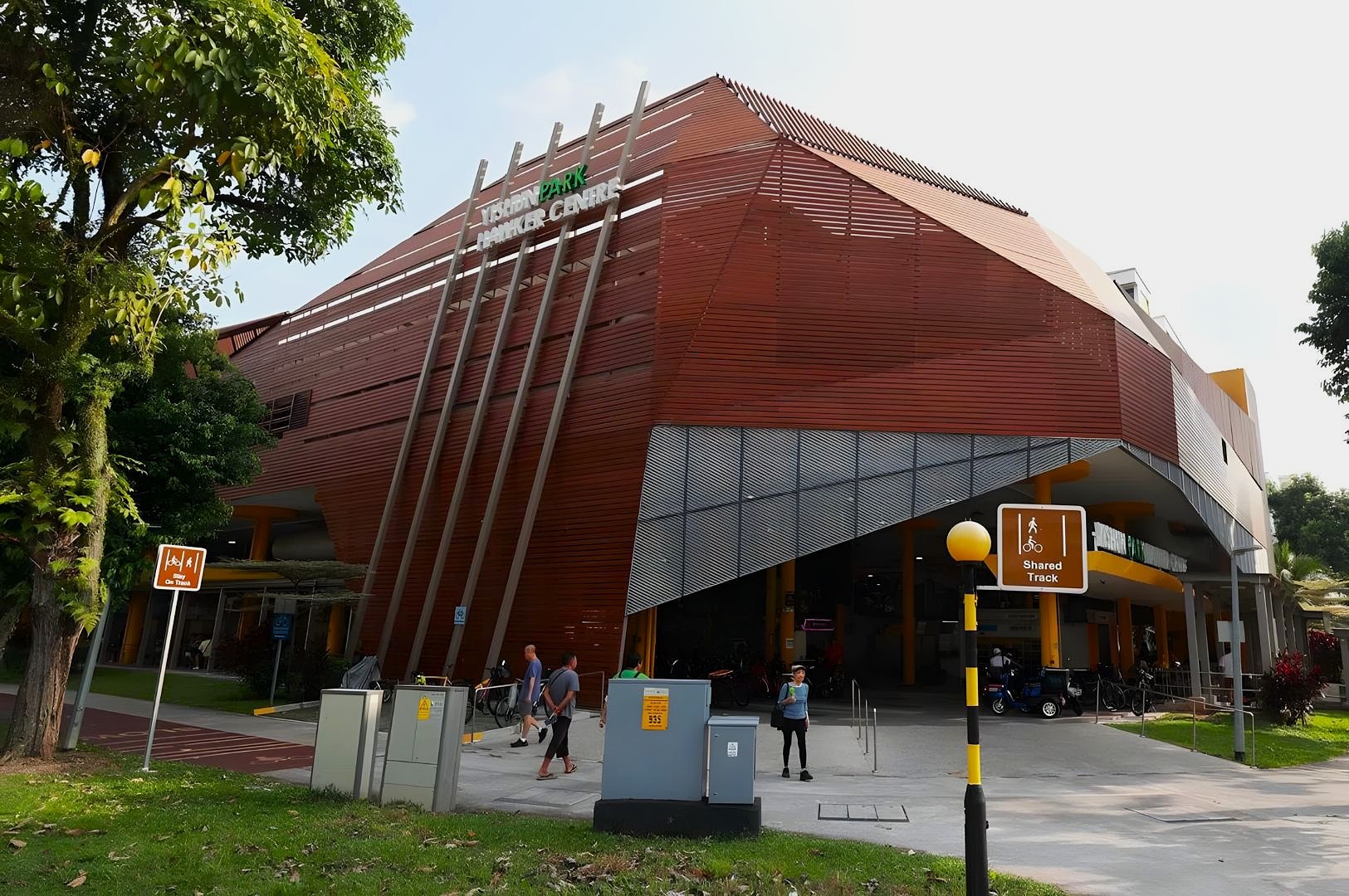 Modern building with red slatted exterior at an urban center, surrounded by trees. People walk on paths, while signs indicate a shared track, creating a lively atmosphere.