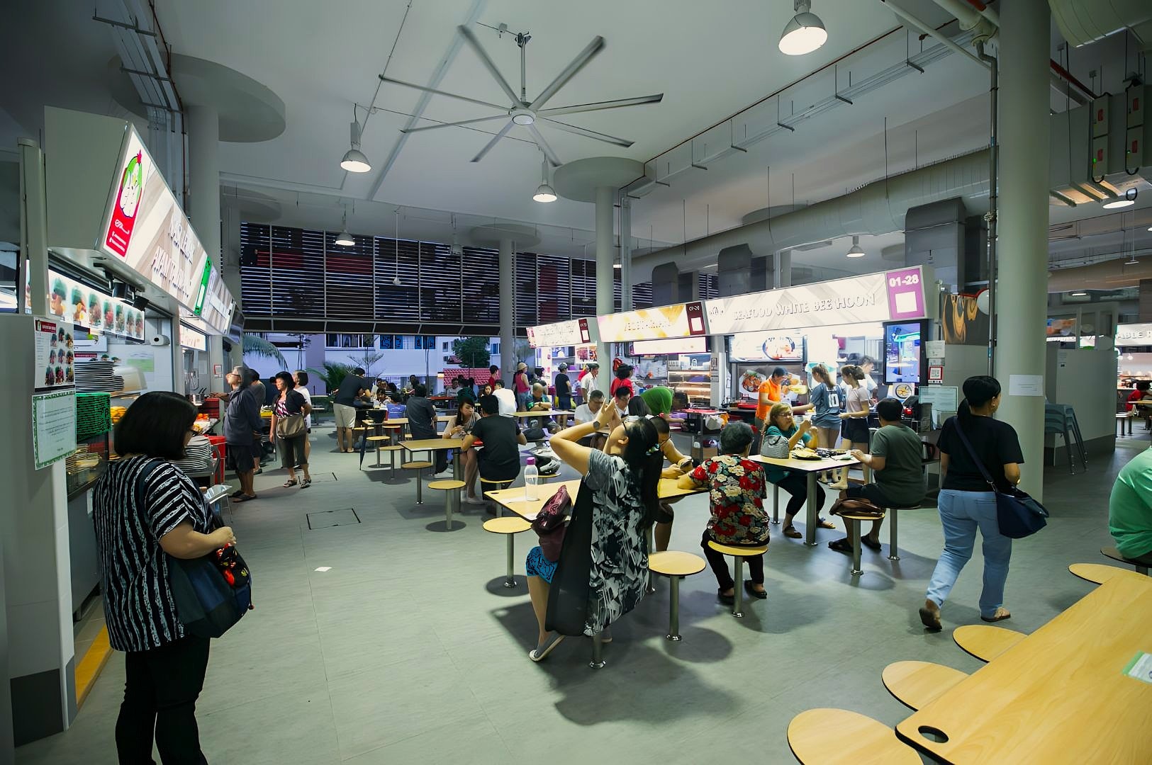 People dine and socialize at a vibrant food court. Bright overhead lights illuminate food stalls and various dishes. The atmosphere is lively and bustling.