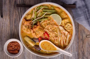 A bowl of spicy laksa noodles is garnished with lime, red chili, green beans, and tofu on a wooden table, next to a small bowl of chili paste.