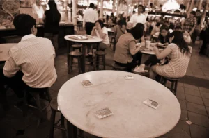 Sepia-toned image of a busy food court with people seated at round tables. Foreground table has four small objects, possibly wallets, indicating reserved seats.