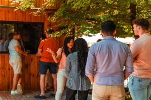 A group of people socializing outdoors near a wooden bar. Two men at the counter, while others chat under sunlit trees. Casual, relaxed atmosphere.