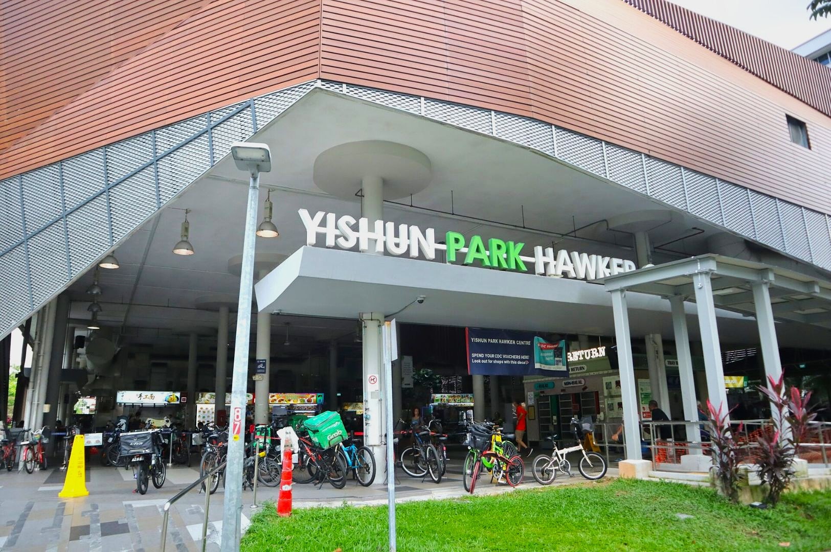 Entrance to Yishun Park Hawker Centre, with parked bicycles and bright signage. People gather inside, conveying a lively, bustling atmosphere.