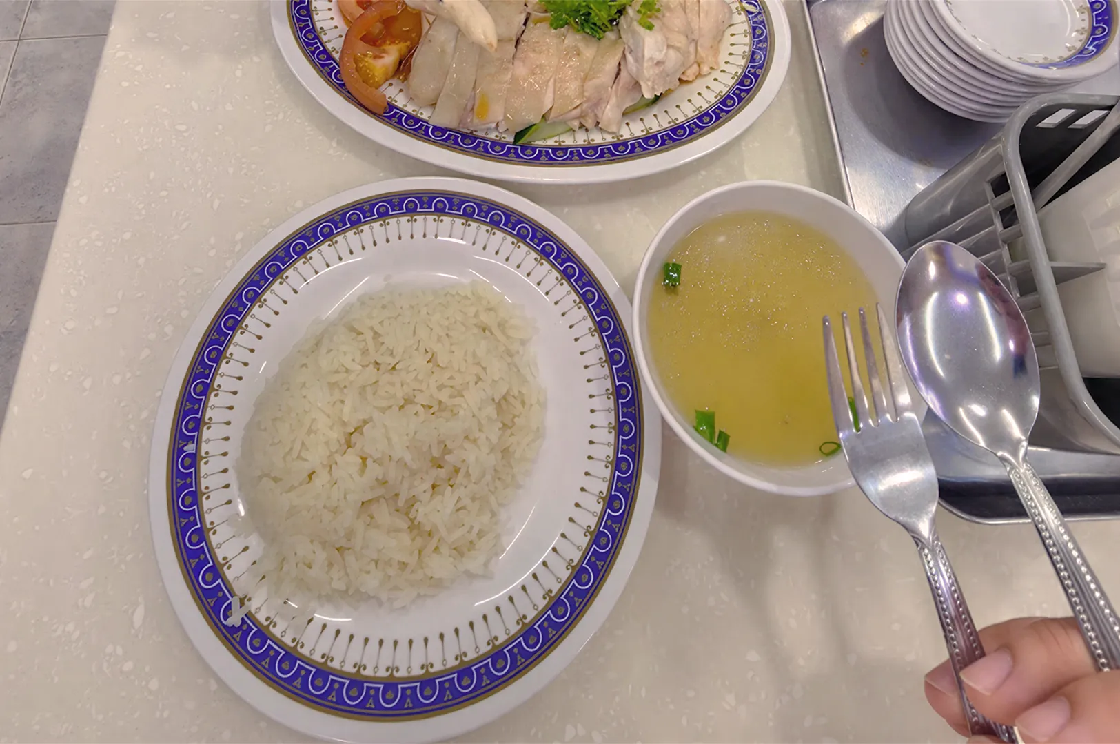 A plate of white rice beside a bowl of clear soup with green onions. Above is a dish of sliced chicken with tomatoes and greens, held with a fork and spoon.