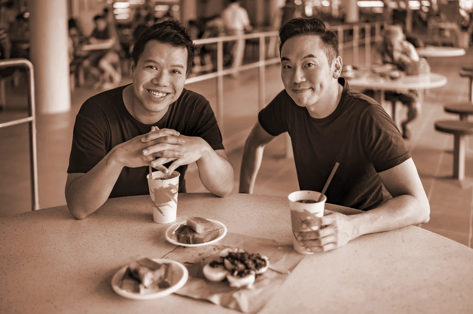 Two people smiling at a food court table with drinks, rainbow-colored desserts, and pastries. The atmosphere is casual and friendly