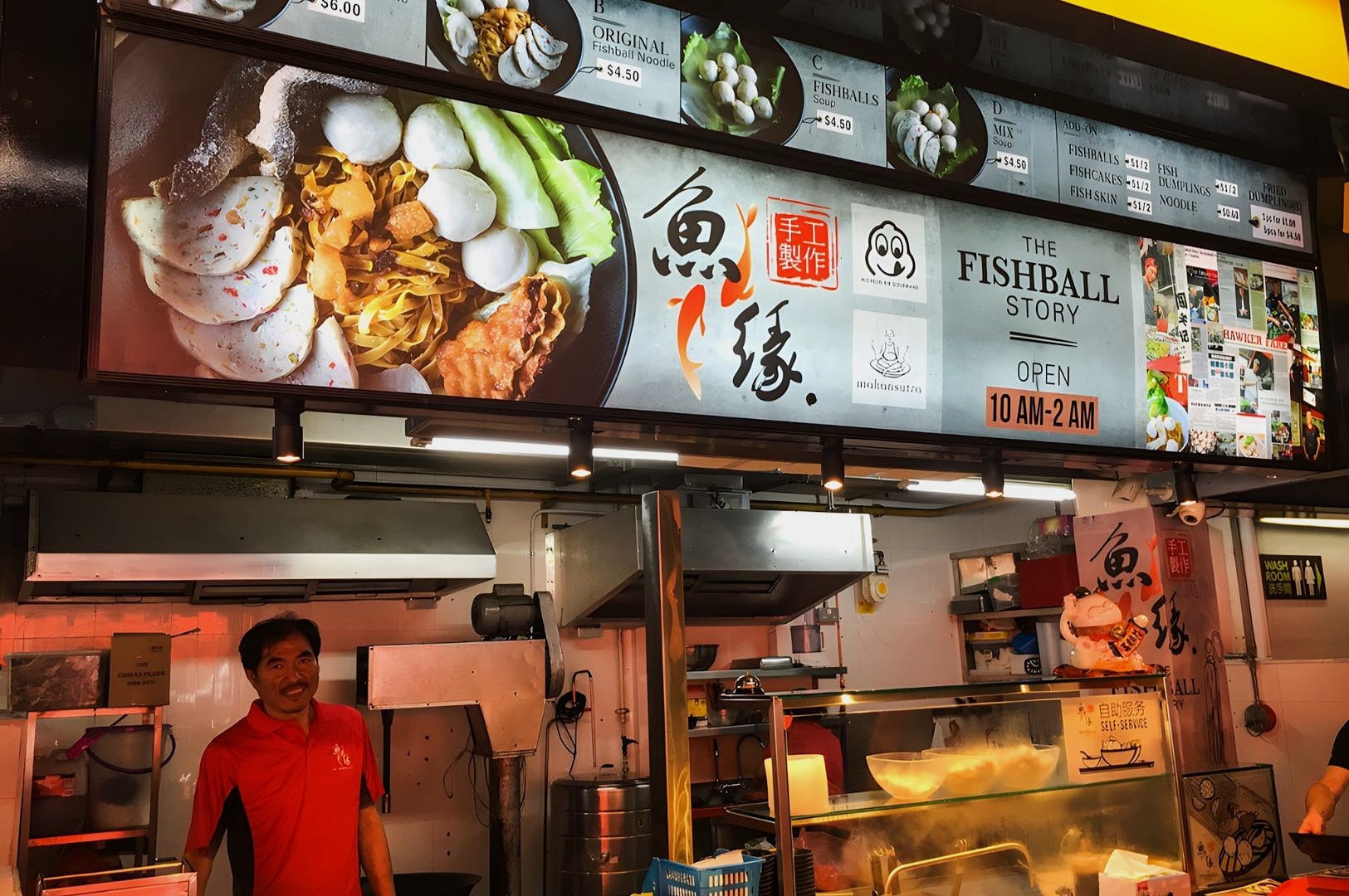 Food stall with colorful signs displaying fishball noodles and menu prices. A smiling vendor in a red shirt stands ready to serve. Bright, inviting ambiance.