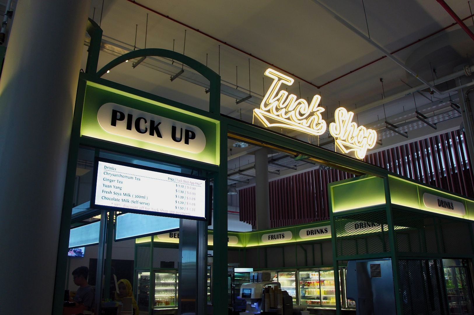 Indoor market stall with a green and black design, labeled "Tuck Shop." Neon signs display "Pick Up" and "Drinks." A menu board lists beverages.