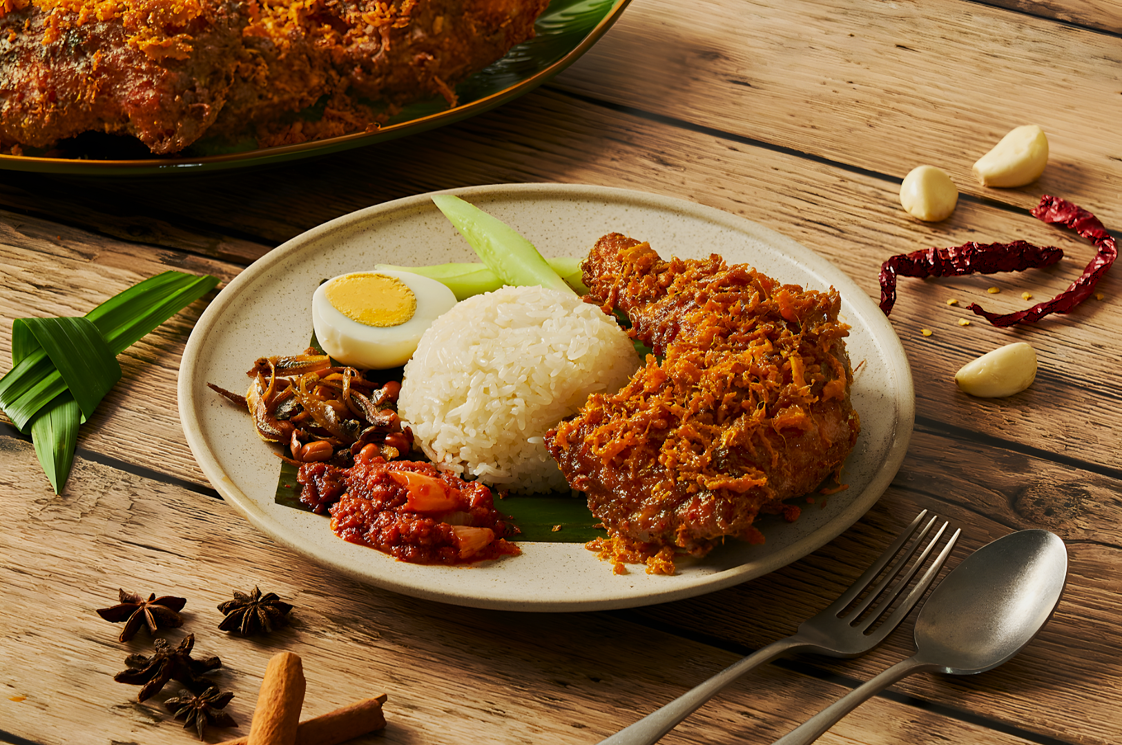 A plate of nasi lemak with coconut rice, fried chicken, half a boiled egg, sambal, anchovies, and cucumber on a rustic wooden table.