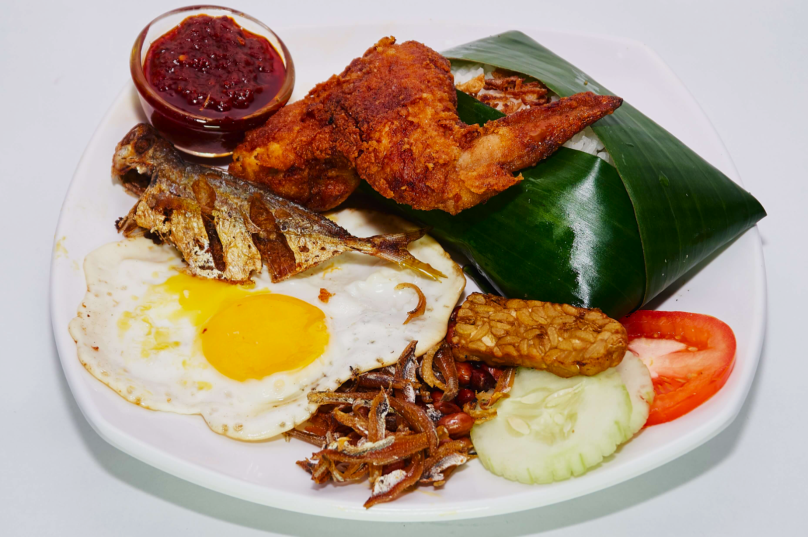 A plate of Malaysian nasi lemak with fried chicken, anchovies, fried egg, sambal, cucumber, tomato, tempeh, and banana leaf-wrapped rice.