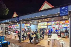 Vibrant nighttime view of Newton Food Centre in Singapore, featuring outdoor hawker stalls with bright signage, patrons dining at tables, and traditional pitched roof architecture.
