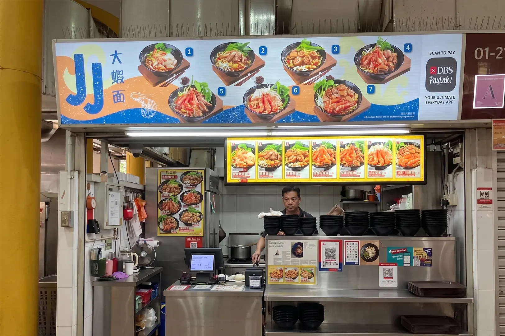 JJ Prawn Noodle hawker stall with a vendor behind the counter and a large illustrated menu signboard.
