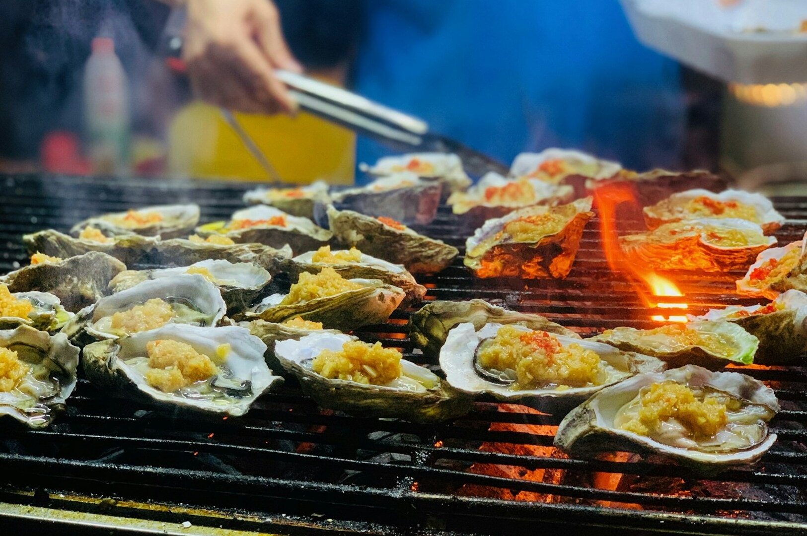 A chef grilling fresh oysters on a grill at Newton Food Centre, with a lively market atmosphere in the background.