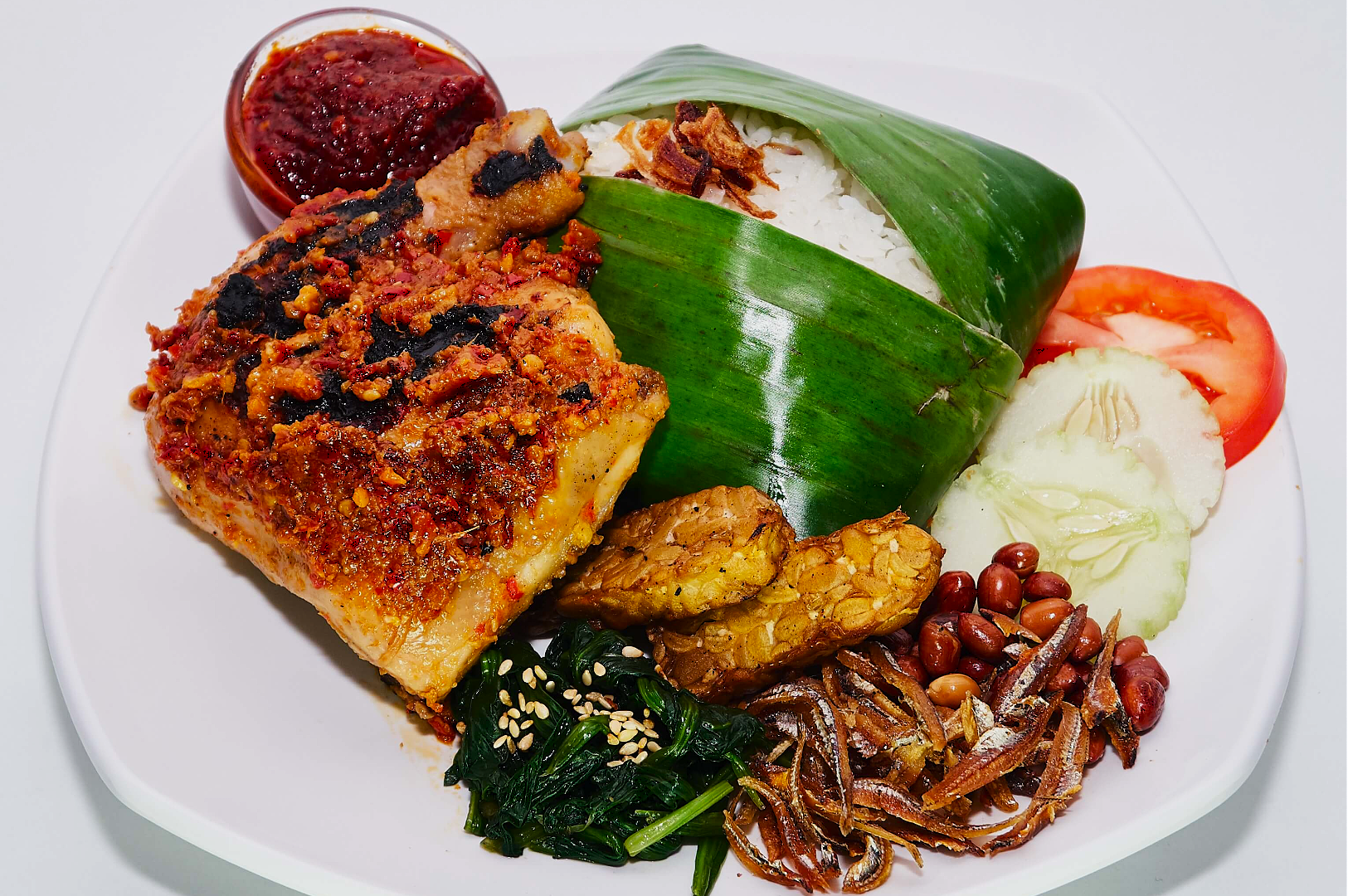A plate of nasi lemak featuring rice in a banana leaf, spicy grilled chicken, sambal, tempeh, spinach, anchovies, peanuts, tomato, and cucumber slices.