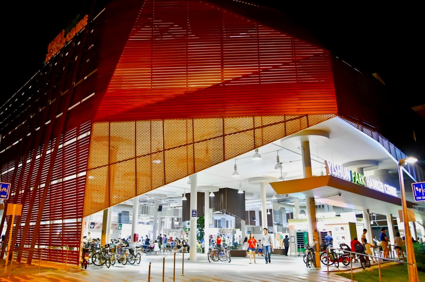 Illuminated at night, this modern building features red lattice architecture. Inside, people gather and bicycles are parked outside, adding a lively vibe.