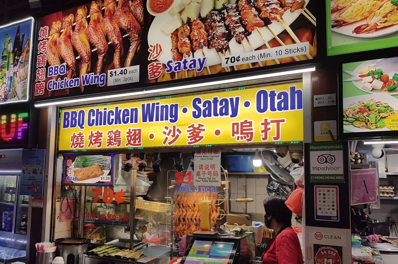 A vibrant food stall displays skewered meats labeled "BBQ Chicken Wing" and "Satay." Bright signage highlights prices and a menu, with a busy vendor in front.