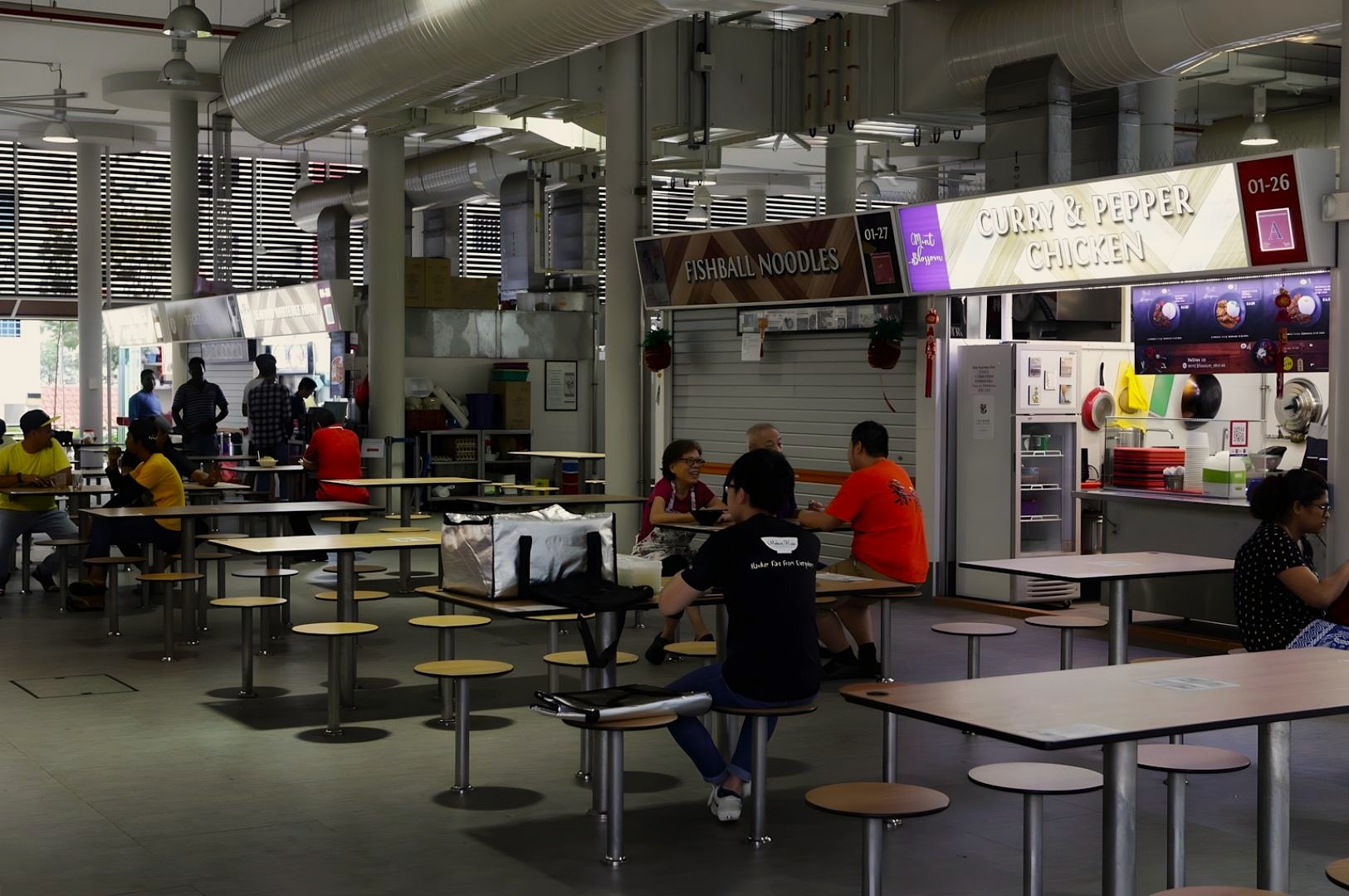 A food court with several patrons seated at tables. Stalls include Curry & Pepper Chicken and Fishball Noodles. The atmosphere is casual and relaxed.