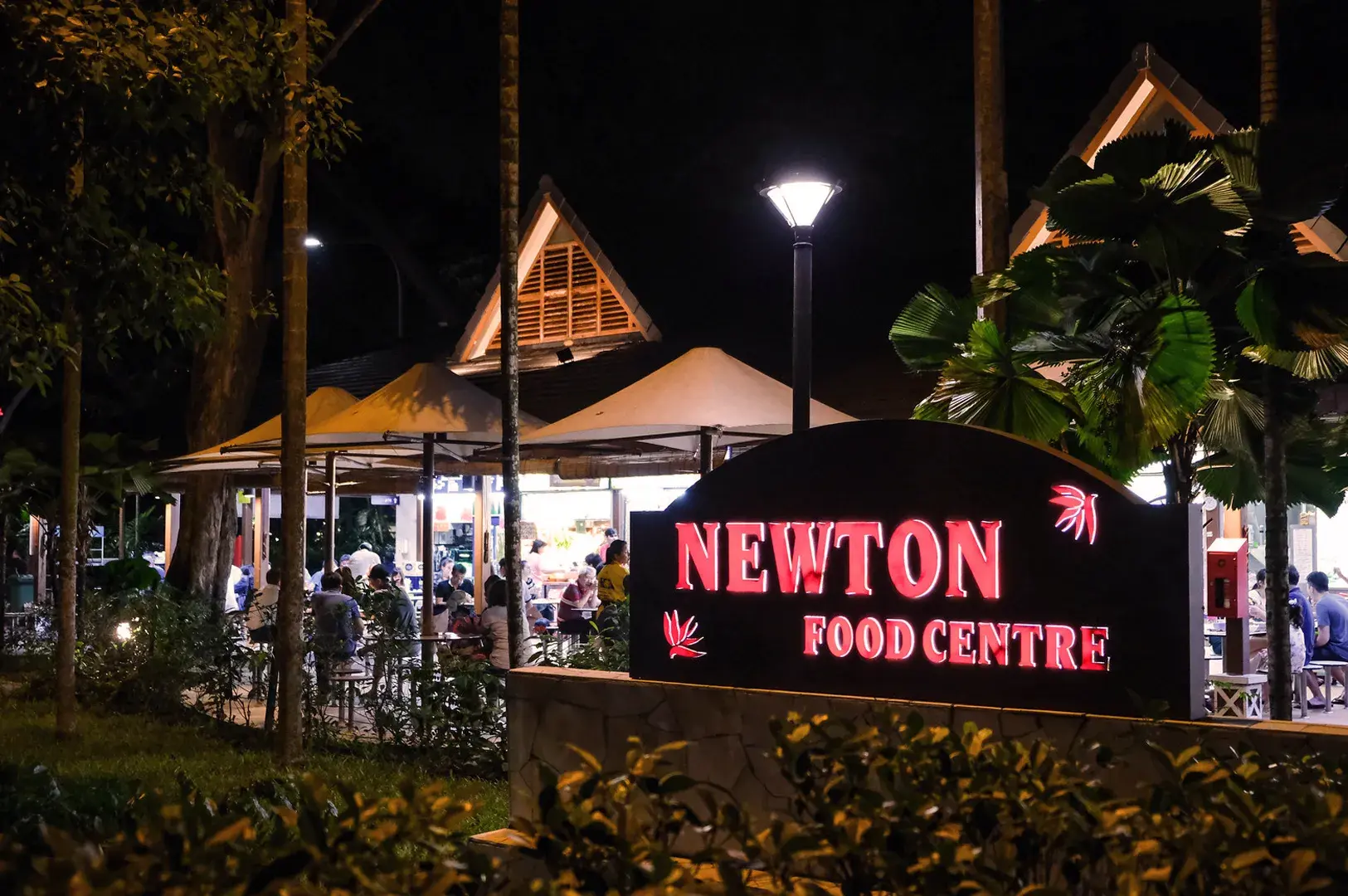 Night scene of Newton Food Centre with a brightly lit red sign in the foreground. Diners sit under a canopy, creating a lively, vibrant atmosphere.