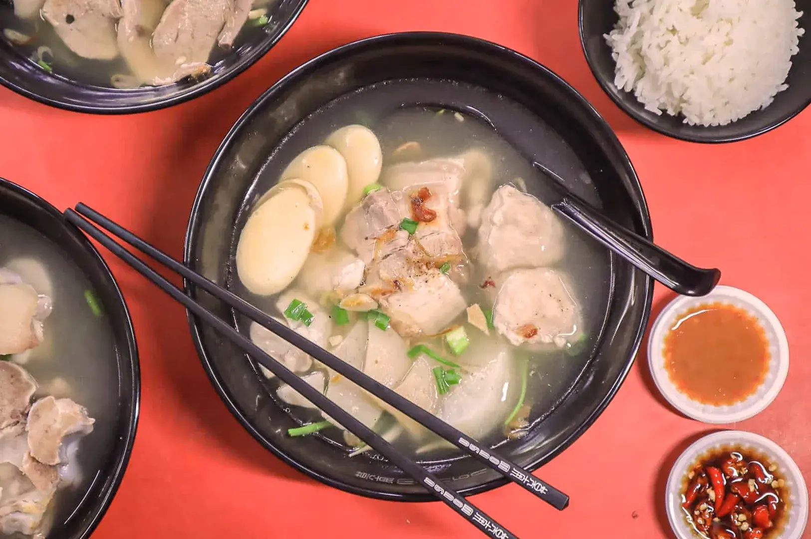 A bowl of soup with chunks of meat, fishcake slices, and green onions on a red table. Chopsticks rest on the bowl. Rice and sauces are on the side.