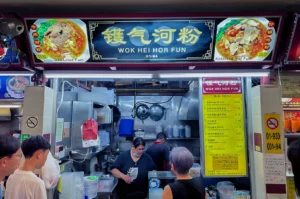 A hawker stall named "Wok Hei Hor Fun" displays its menu. People are ordering food while the cooks prepare dishes in a bustling, vibrant setting.