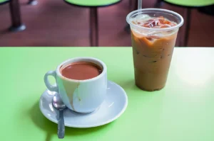 A white cup of hot coffee on a saucer with a spoon and a plastic cup of iced coffee with a straw sit on a bright green table.