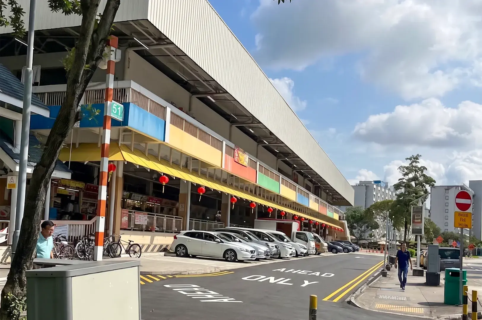 Street view of a multi-story building featuring colorful awnings and red lanterns. Cars are parked in front along a road marked "AHEAD ONLY," with pedestrians walking on the sidewalk under a sunny sky.