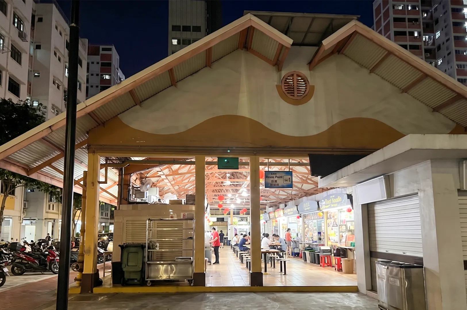 A night view of a brightly lit food court interior with several food stalls and people dining at tables under a large roof.