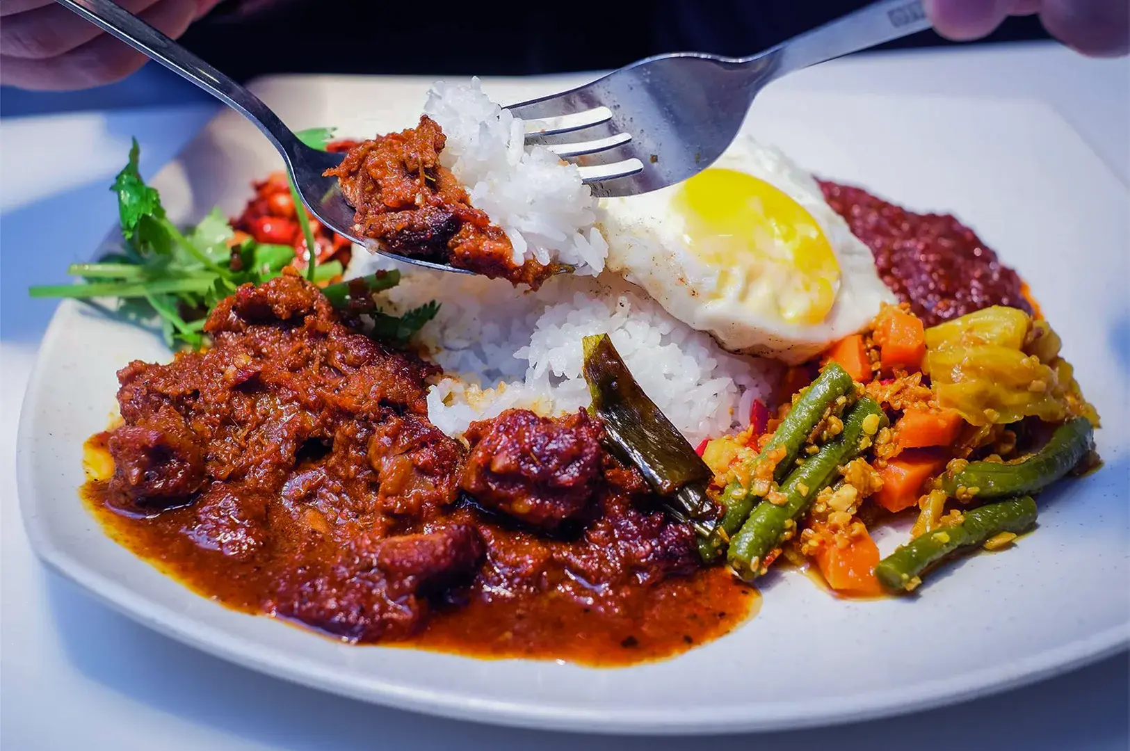 A plate of nasi lemak with coconut rice, fried egg, spicy meat curry, vegetables, sambal, and a fork scooping rice, surrounded by greens.