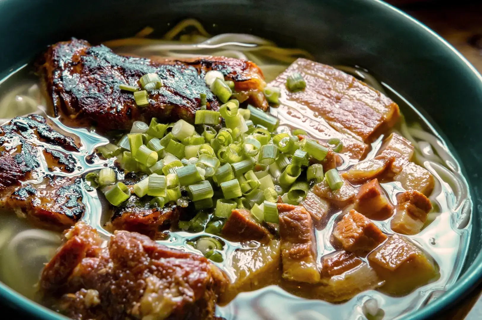 A close-up of a steaming bowl of ramen. Grilled pork, diced green onions, and broth-covered noodles create a savory and comforting meal.