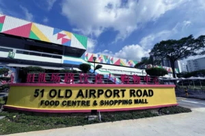 Sign reading '51 Old Airport Road Food Centre & Shopping Mall' in front of a colorful building under a partly cloudy sky. Lush greenery surrounds the area."