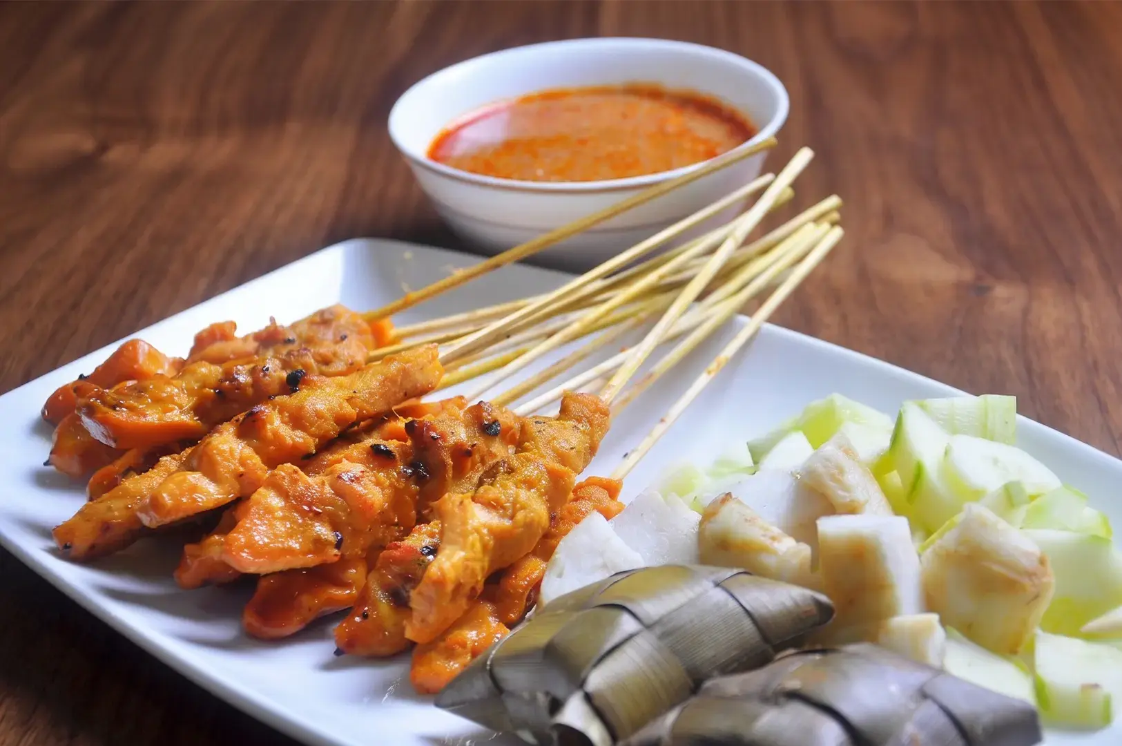 Grilled chicken satay skewers on a white plate, served with ketupat, sliced cucumbers, and a side of peanut dipping sauce.