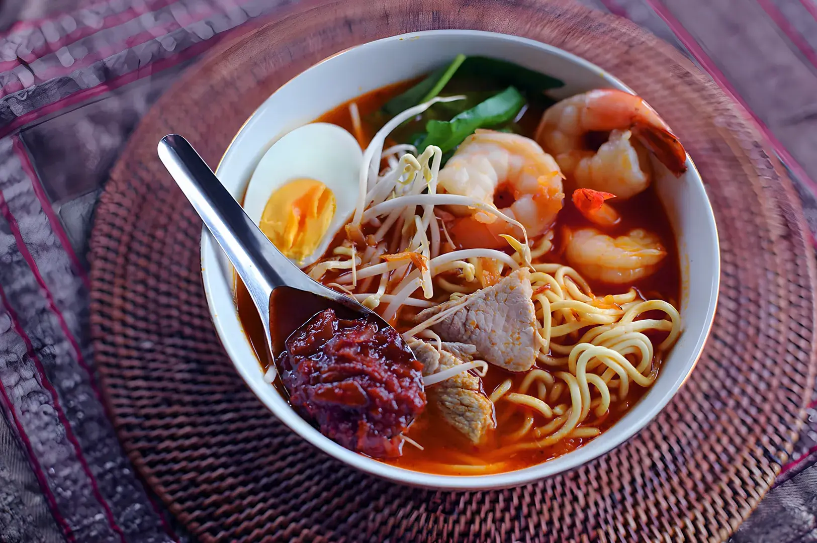 A bowl of spicy noodle soup with shrimp, sliced egg, bean sprouts, meat, and a spoonful of chili paste, set on a woven mat, conveying warmth.