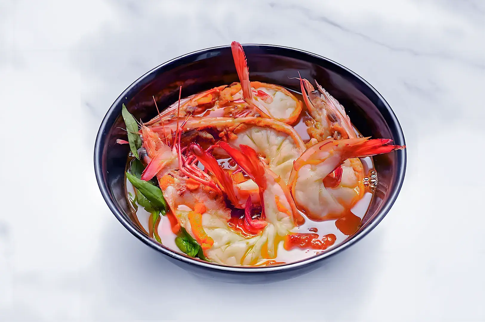 A black bowl of spicy shrimp wonton soup, featuring large shrimp, vegetables, and wontons in a red chili-infused broth. The dish looks vibrant and appetizing.