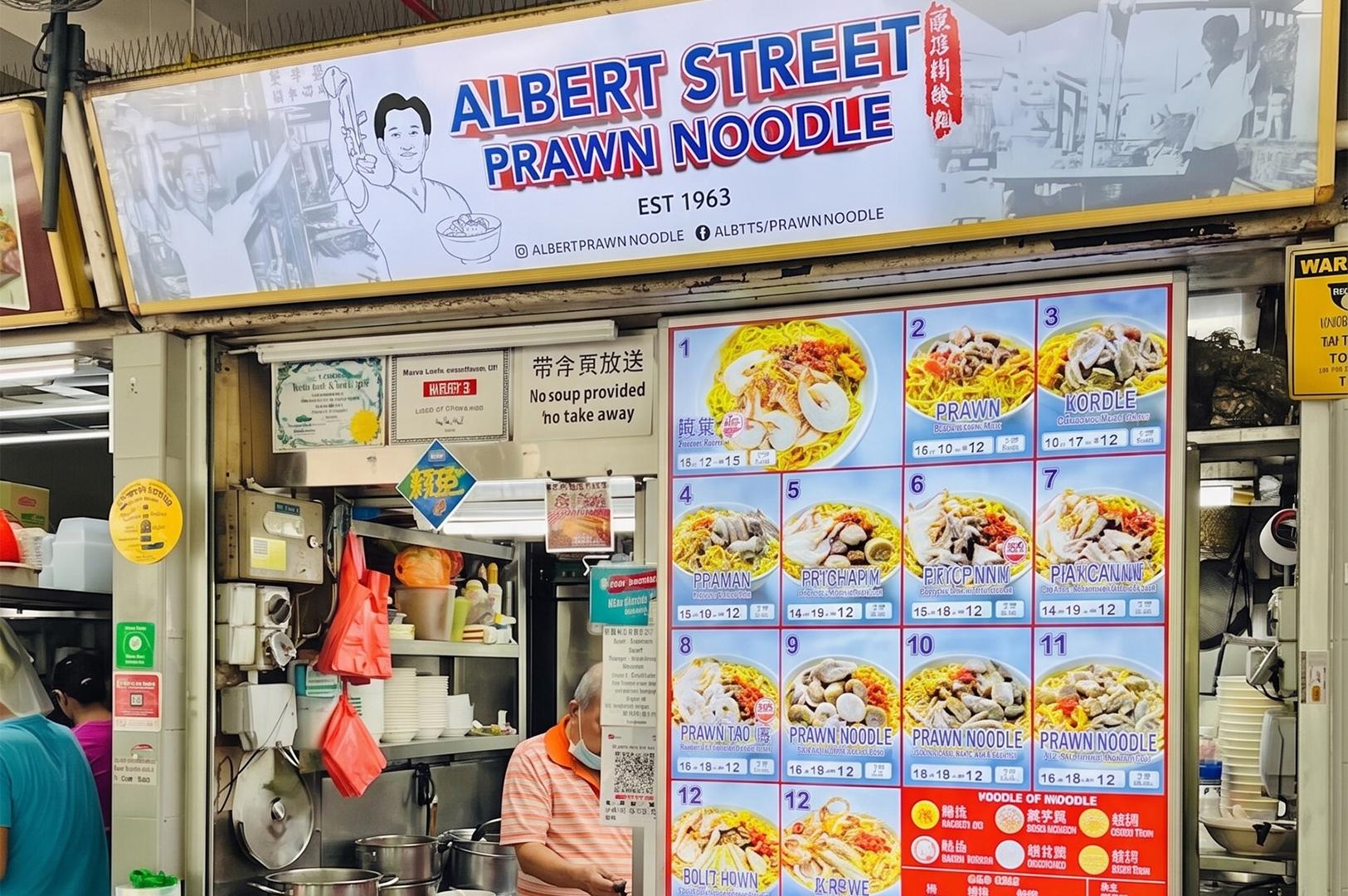 A hawker stall named "Albert Street Prawn Noodle" with a large illustrated menu board showing various noodle dishes and an elderly man working behind the counter.