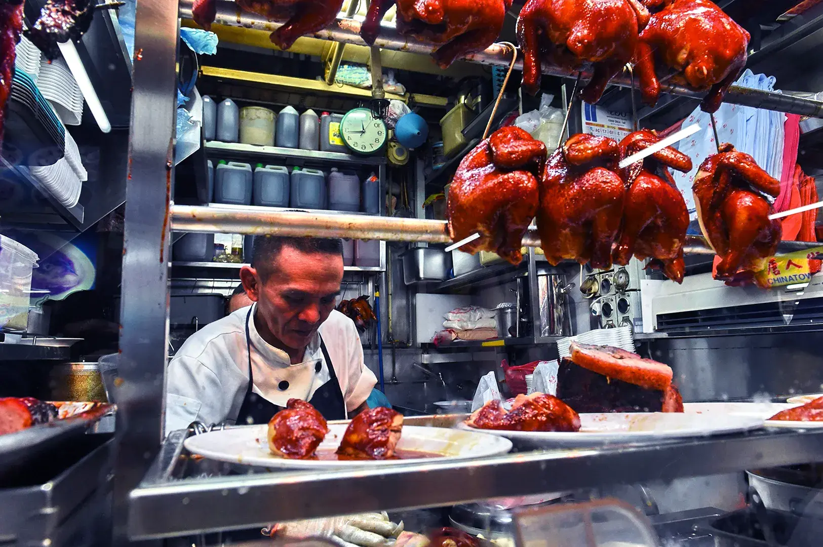 A street food vendor in a small kitchen, preparing skewered roasted birds. The scene is busy and vibrant, with a clock and stacked containers in the background.