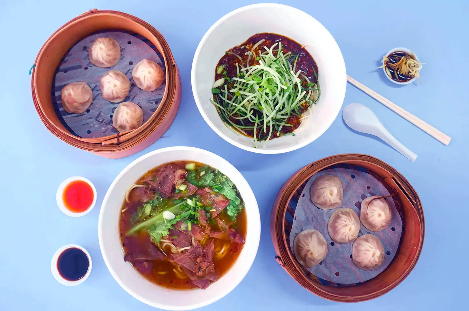 Overhead view of two baskets of dumplings and two noodle dishes on a blue surface. Small bowls of sauce, chopsticks, and a spoon complete the vibrant scene.